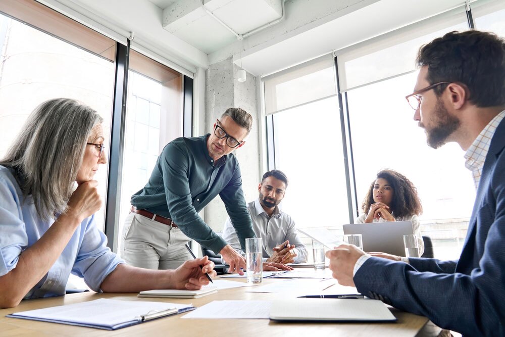 Group of professionals gathered around a conference table in a modern office – one person standing and gesturing while others listen attentively with notebooks and laptops open; scene reflects teamwork, discussion, and strategic collaboration.