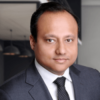 Professional man wearing a gray suit, white dress shirt, and black patterned tie; posing confidently in a modern office environment with glass walls and pendant lighting in the background.